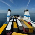Aerial view of container trucks transported on a large ferry across a calm ocean under a clear blue sky.