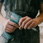 Crop anonymous mechanic in uniform with wireless drill standing in flat during repair works